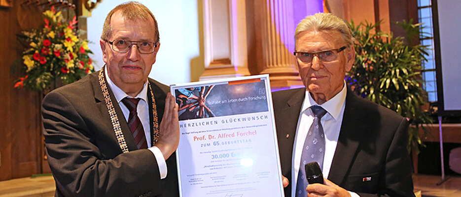 Dr. Kurt Eckernkamp (rechts) überreicht Unipräsident Alfred Forchel in der Neubaukirche die Urkunde mit dem Förderbescheid. (Foto: Rudi Merkl)