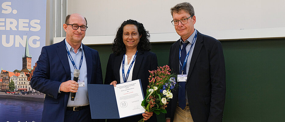Die diesjährige DGHM-Hauptpreisträgerin Cynthia Sharma bei der Preisverleihung mit DGHM-Präsident Prof. Jan Buer (r) und Prof. Klaus Pfeffer, dem Vorsitzenden der DGHM-Stiftung.
